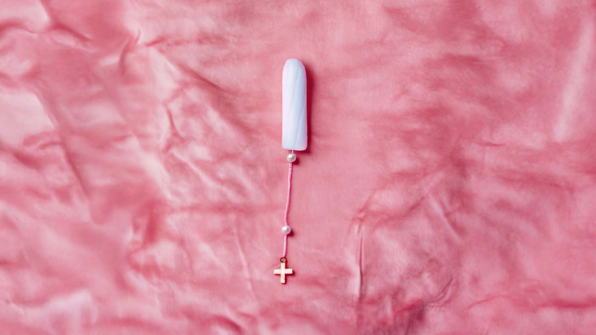 Tampon mit Kreuzanhänger auf rosa Stoff, Mischung aus Midjourney und Photoshop. O.B.-Damenhygieneartikel, feminine Hygiene, deaex 2025, designaustria