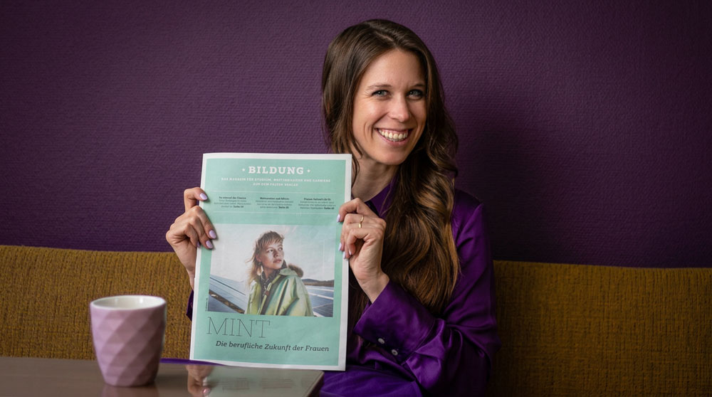 Smiling woman in a purple blouse sits on a purple wall, holding a mint magazine titled 'MINT' about women's future.