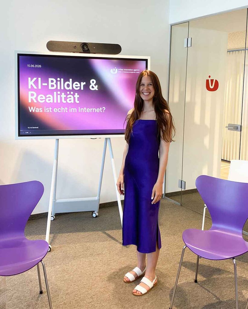 Tiny Bernhard von deaex in a royal purple dress stands beside a presentation screen in a conference room, smiling at the camera.