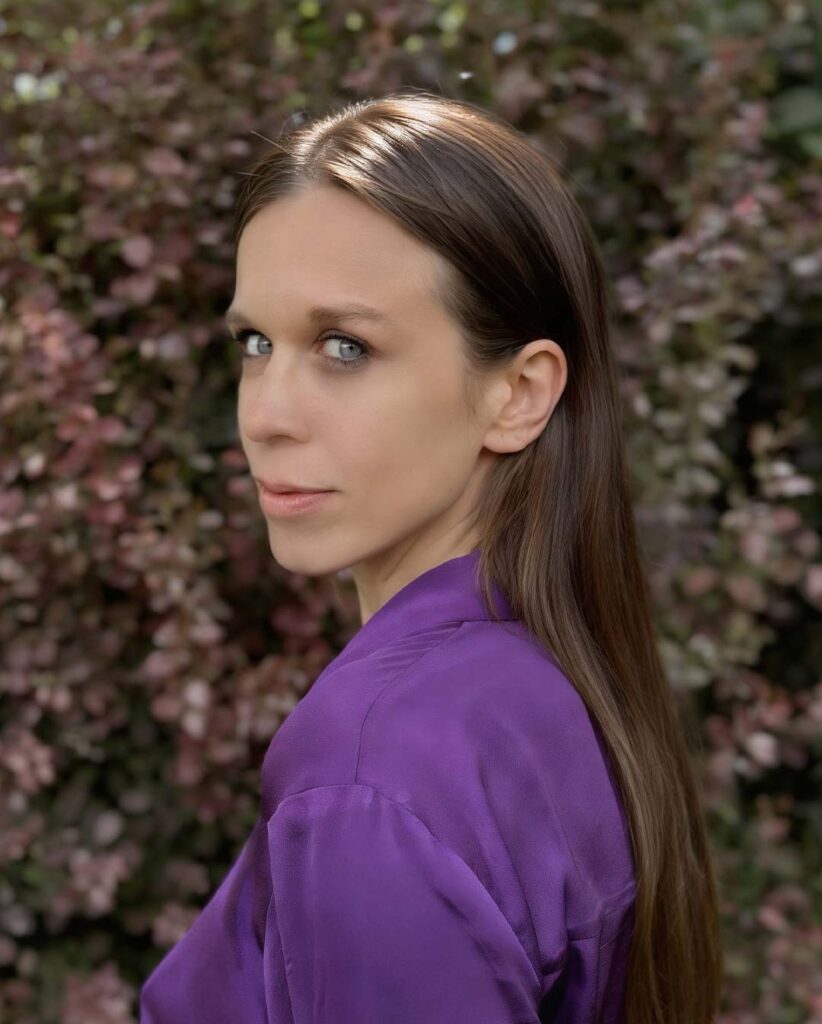 Tiny Bernhard von deaex of a woman with long brown hair wearing a purple blouse, standing in front of pink flowering bushes.
