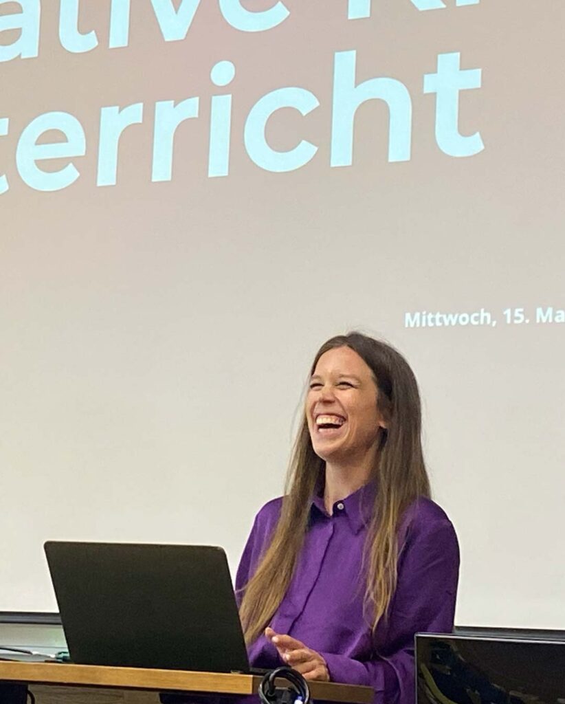 Tiny Bernhard von deaex in a purple shirt laughing during a presentation, standing at a desk with a laptop as a projection fills the background.