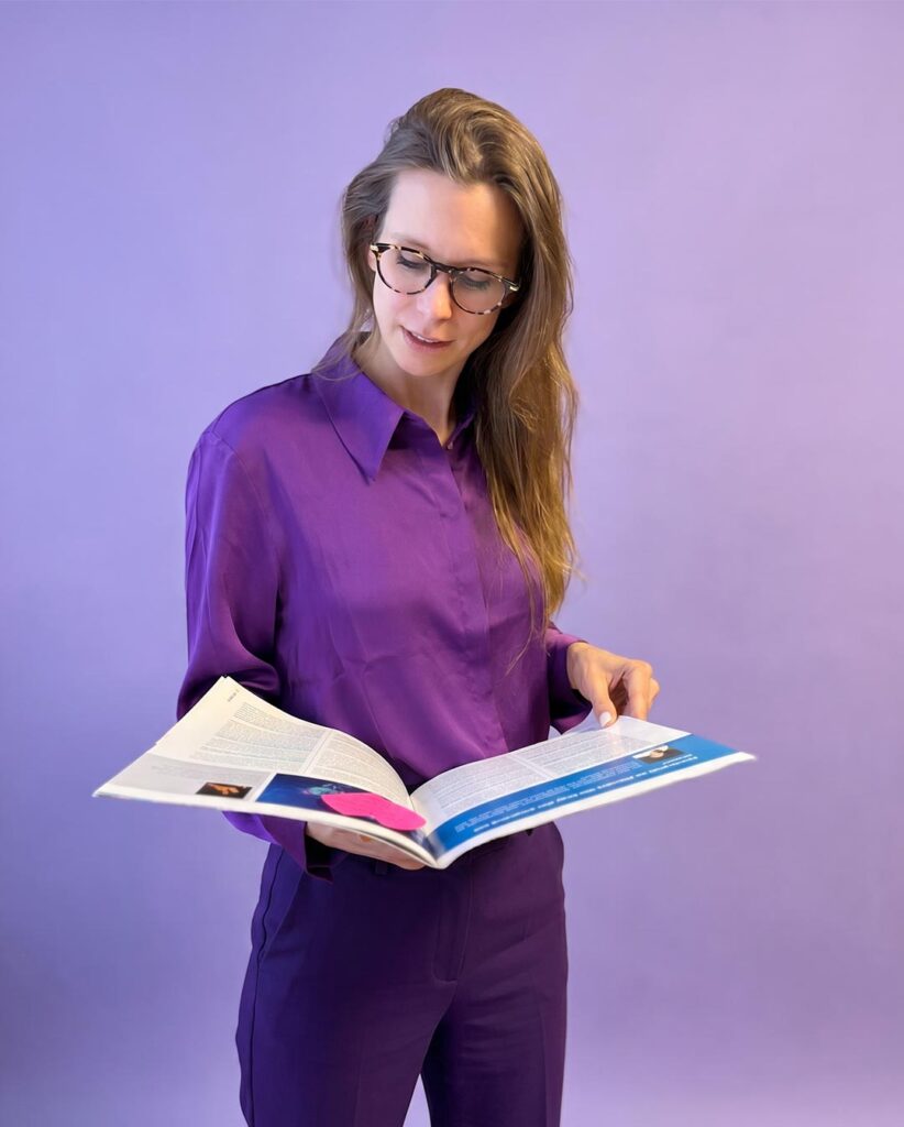 Tiny Bernhard von deaex in a purple blouse and pants reads a magazine against a purple background, wearing glasses.