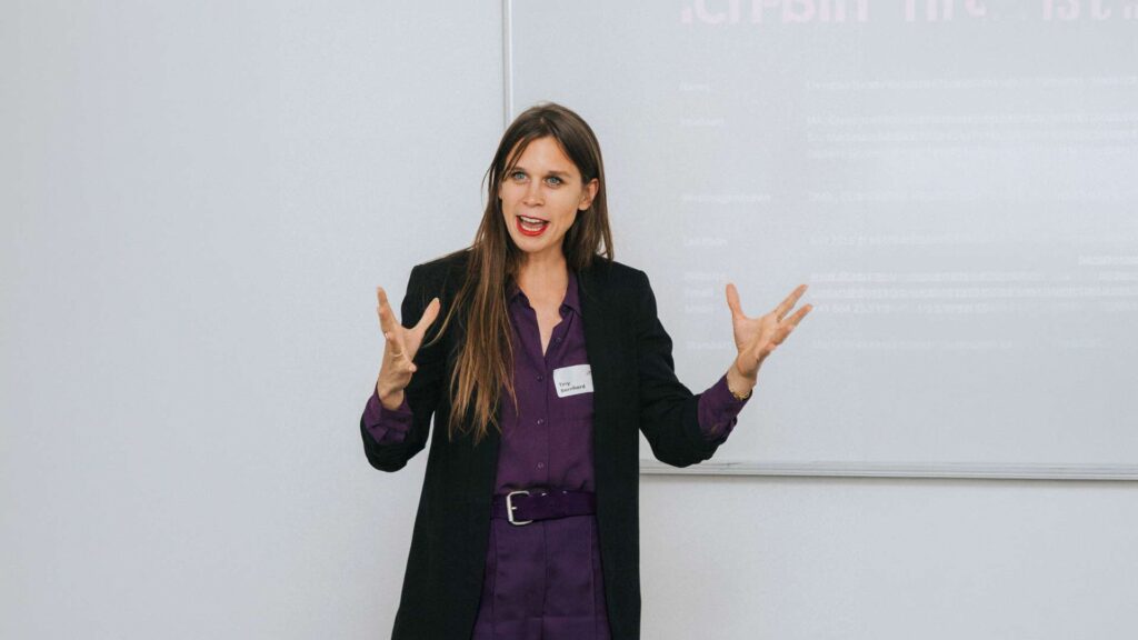 Woman in a purple shirt and black blazer giving a presentation, hands raised as she speaks in front of a whiteboard.