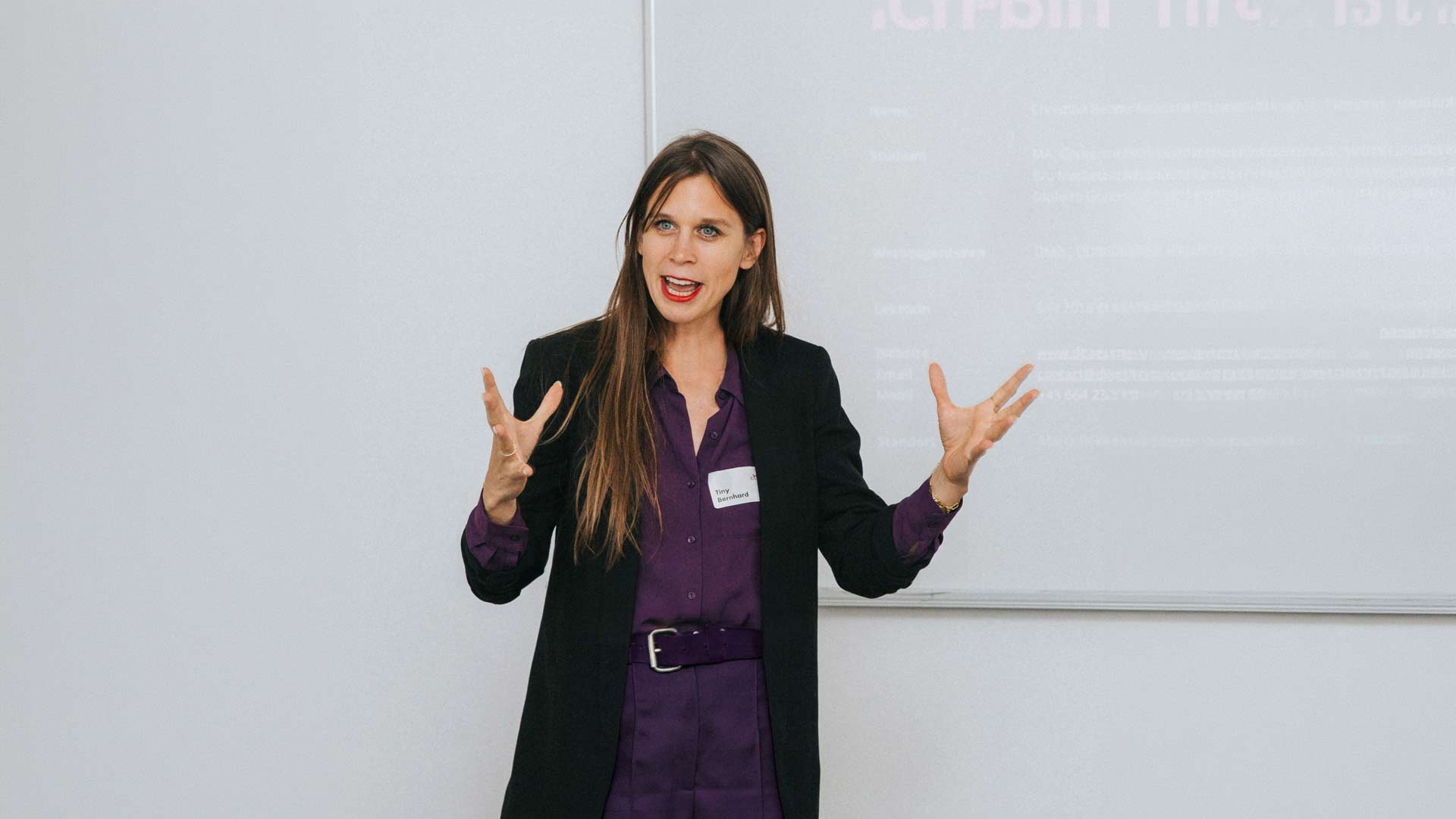Woman in a purple shirt and black blazer giving a presentation, hands raised as she speaks in front of a whiteboard.