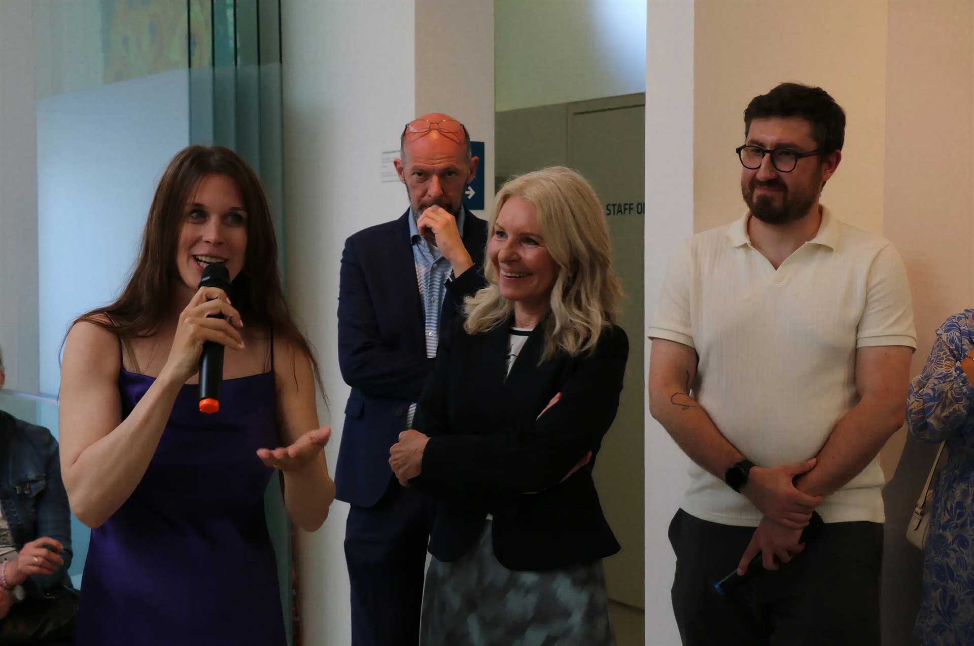 Woman in a purple dress speaks into a microphone at a panel or group event, gesturing with her hands, with three people watching.