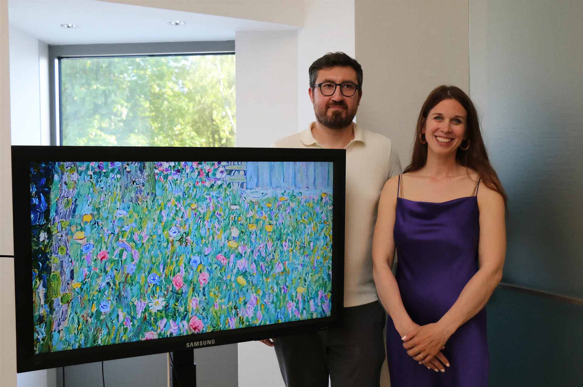 Man with glasses and a woman in a purple dress stand beside a large Samsung monitor displaying a colorful abstract painting in an office.