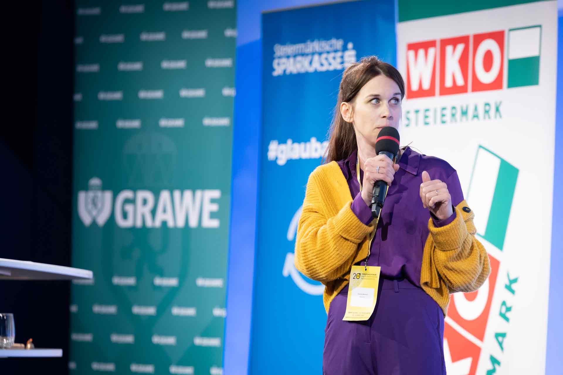 Female presenter on stage speaking into a microphone, wearing a purple outfit and yellow cardigan, with conference banners in the background.