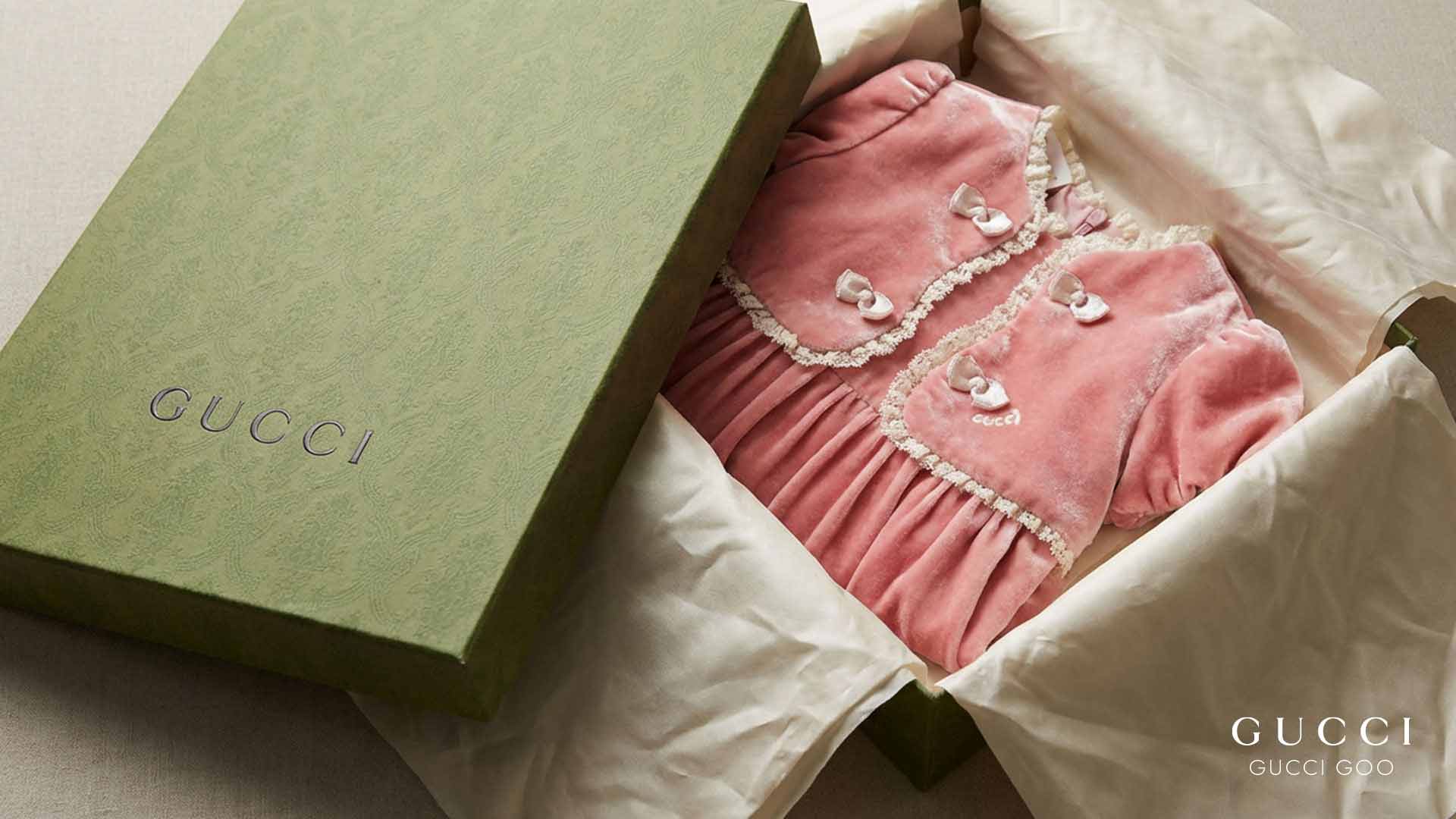 Pink velvet Gucci dress with white lace trim and bows, shown in an open green gift box with tissue paper.