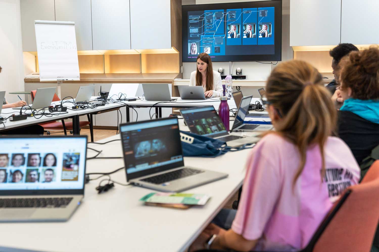 Instructor leads a laptop workshop at the front of a classroom as students work on laptops and a large data screen is shown behind.
