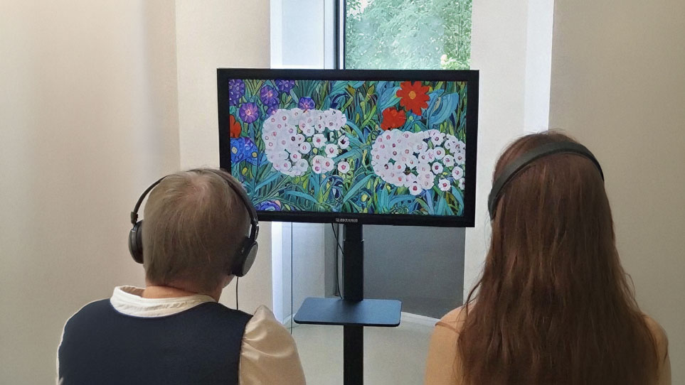 Two people with headphones stand in front of a monitor displaying a bright, colorful floral painting. A window is visible behind them.