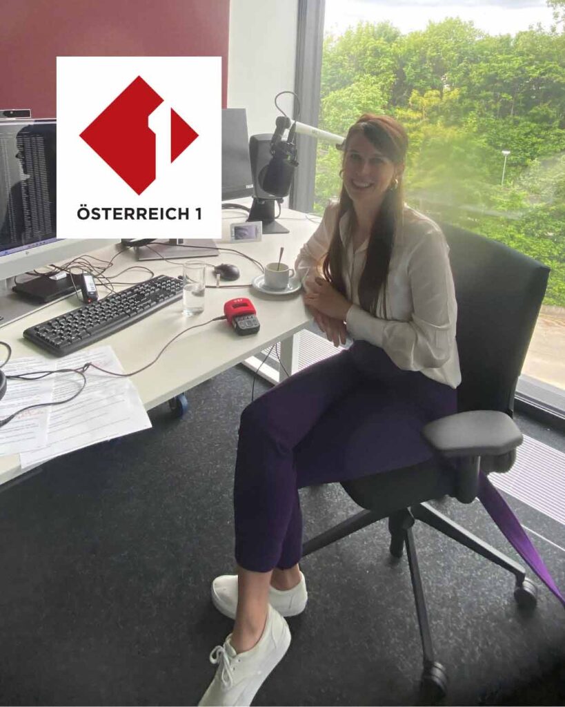 Woman radio host sitting at a desk in a studio, smiling, with a microphone, computer keyboard, and a coffee cup nearby; Österreich 1 logo overlay on the left.